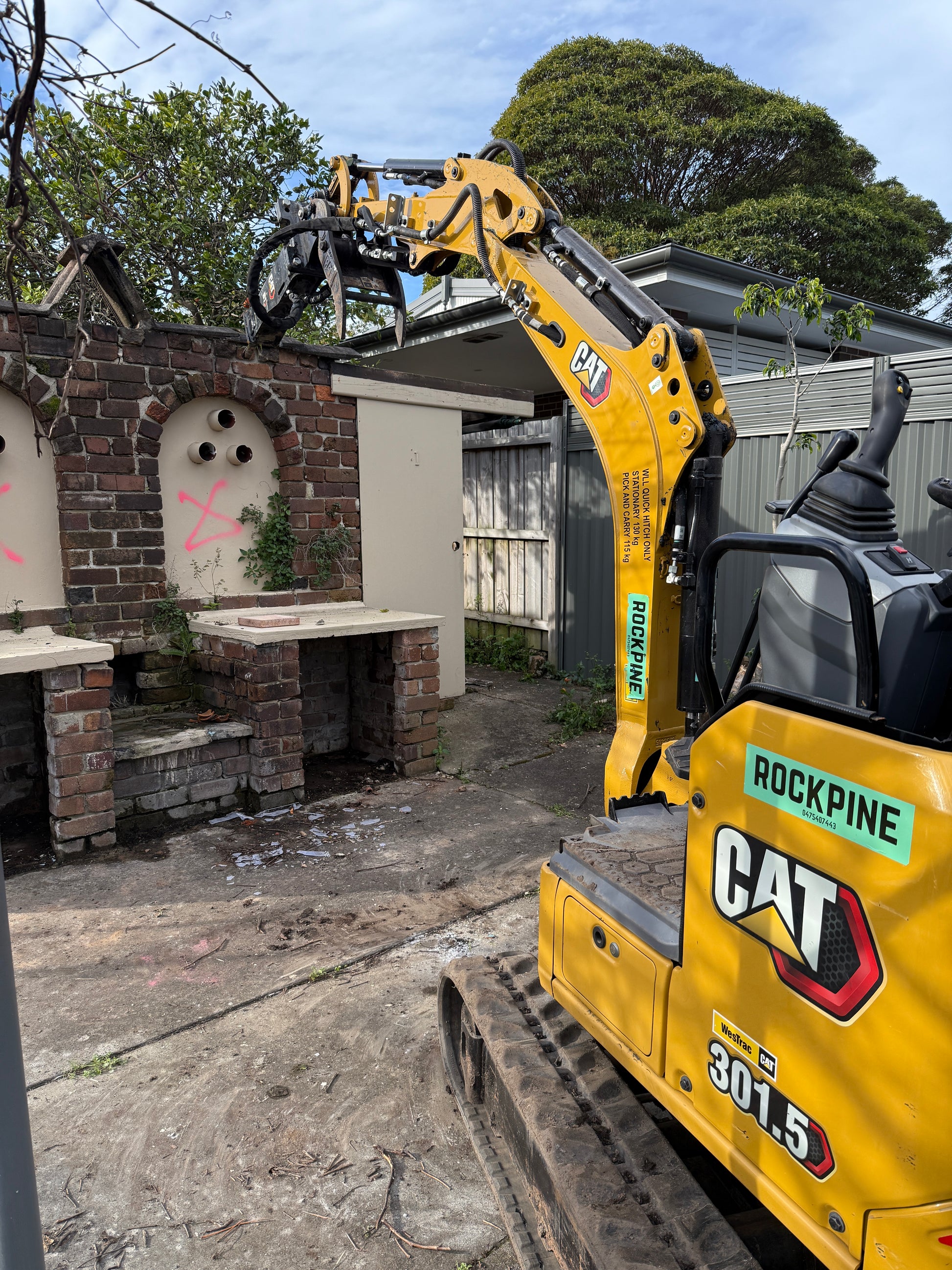 Cat excavator performing demolition work on a construction site with trees and building in the background.